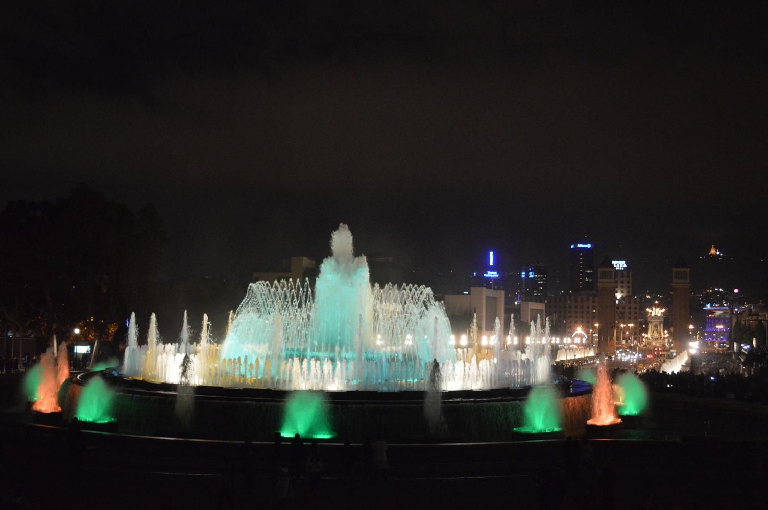 Magic Fountain of Montjuïc in Barcelona illuminated at night with vibrant multicolored water jets, city buildings and towers in the background, and a lively urban atmosphere.